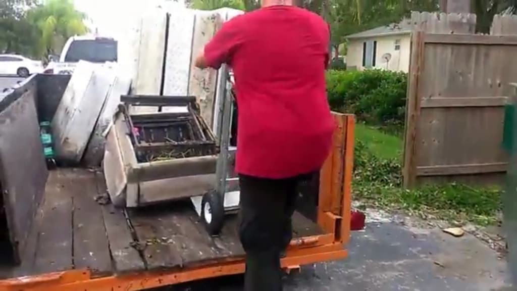 Hauler loading a bulky item onto a flatbed trailer at the curb outside a suburban home