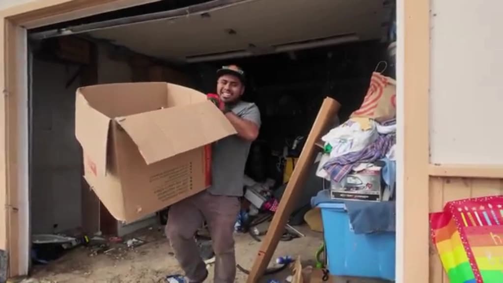 Crew member carrying a large cardboard box out of a garage filled with stored belongings