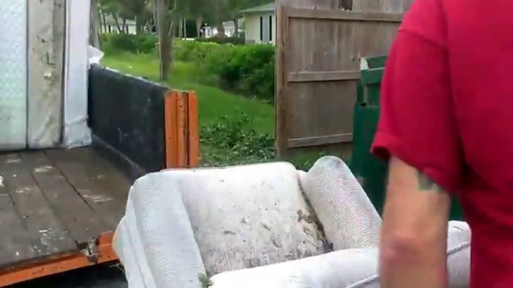 Weathered upholstered armchair being lifted onto a trailer beside a back fence