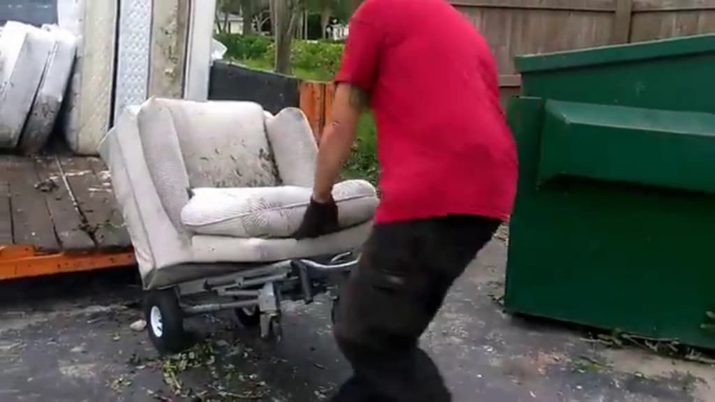 Crew member wheeling a worn armchair out on a hand truck toward a waste container